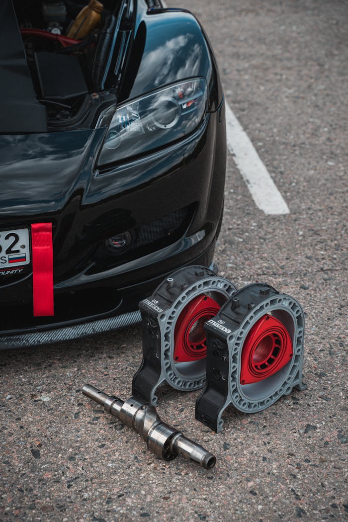 Detailed view of car engine parts on asphalt near a black sports car, showcasing automotive maintenance.